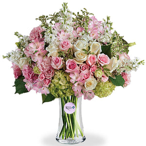 A floral arrangement featuring green hydrangea, pink roses, white roses, pink spray roses, light pink alstroemeria, pink lisianthus, and white stock in various shades of pink, green, and white tones, presented in a clear glass vase.