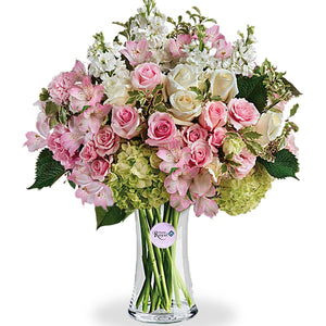 A floral arrangement featuring green hydrangea, pink roses, white roses, pink spray roses, light pink alstroemeria, pink lisianthus, and white stock in various shades of pink, green, and white tones, presented in a clear glass vase.